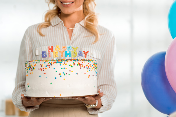 przycięty widok uśmiechniętej kobiety trzymającej tort urodzinowy z kolorowymi świeczkami i szczęśliwym napisem urodzinowym - Zdjęcie, obraz