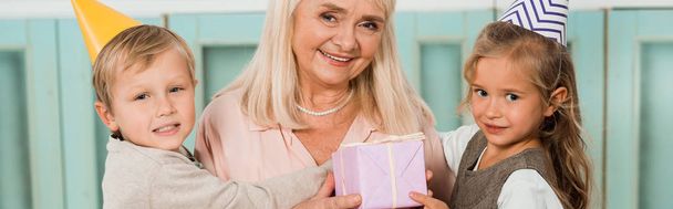 πανοραμική λήψη των εγγονιών αγκαλιάζει ευτυχισμένη γιαγιά κρατώντας κουτί δώρου και χαμογελώντας στην κάμερα - Φωτογραφία, εικόνα