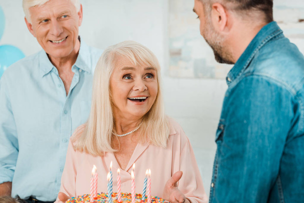 šťastný senior žena drží narozeninový dort, zatímco stojí v blízkosti manžela a dospělého syna - Fotografie, Obrázek