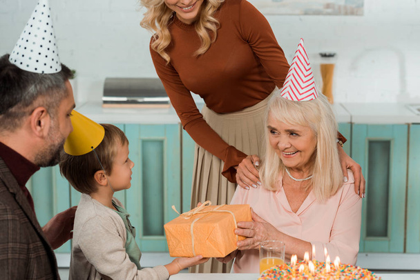 обрізаний вид жінки торкається плечей щасливої бабусі, беручи подарункову коробку від онука
 - Фото, зображення