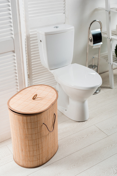 εσωτερικό από λευκό μοντέρνο μπάνιο με λεκάνη τουαλέτας κοντά σε πτυσσόμενη οθόνη, καλάθι πλυντηρίων, βούρτσα τουαλέτας - Φωτογραφία, εικόνα