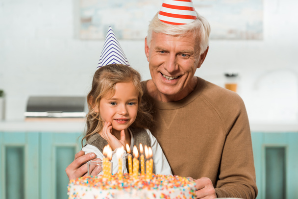 gelukkig senior man met schattige kleindochter op zoek naar verjaardagstaart met brandende kaarsen - Foto, afbeelding