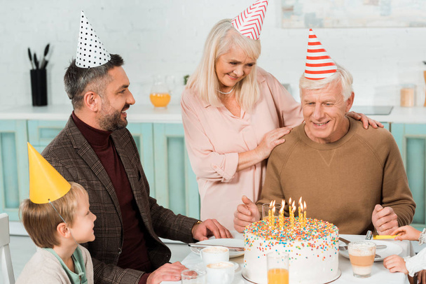 Seniorin berührt Schultern eines glücklichen Mannes, der neben Geburtstagstorte mit Familie sitzt - Foto, Bild