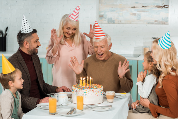 взволнованный пожилой человек смотрит на торт с горящими свечами, сидя за обслуживаемым столом рядом с семьей
 - Фото, изображение