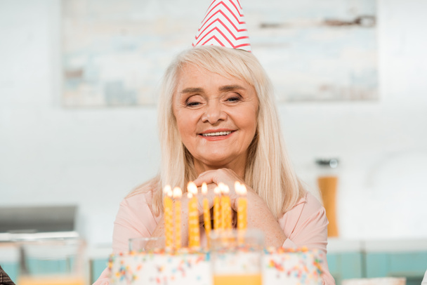 happy senior woman in party cap looking at birthday cake with burning candles - Photo, Image