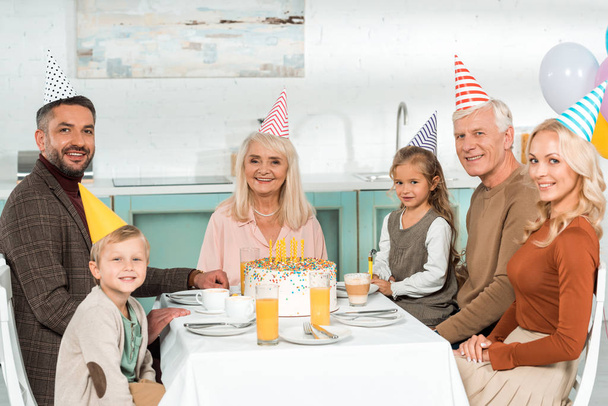 glückliche Familie blickt in die Kamera, während sie am Küchentisch mit Geburtstagstorte sitzt - Foto, Bild