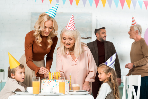 パーティーキャップで家族の近くの誕生日ケーキにろうそくを置く幸せな女性 - 写真・画像