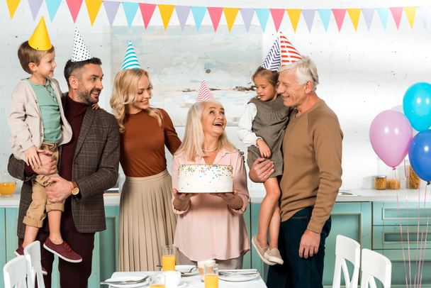 happy senior woman holding birthday cake near cheerful family in party hats - Photo, Image