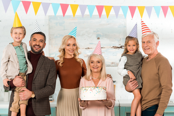 Glückliche Seniorin mit Geburtstagstorte neben Familie in Partyhüten lächelt in die Kamera - Foto, Bild