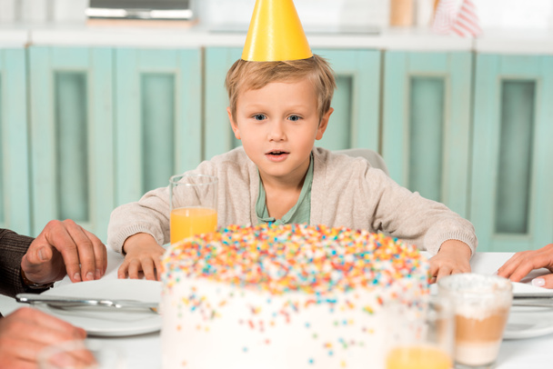 selektivní zaměření užaslého chlapce v čepici strany při pohledu na narozeninový dort na kuchyňském stole - Fotografie, Obrázek