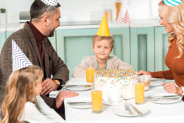 cute kid looking at birthday cake while sitting at served table near parents and sister - Photo, Image