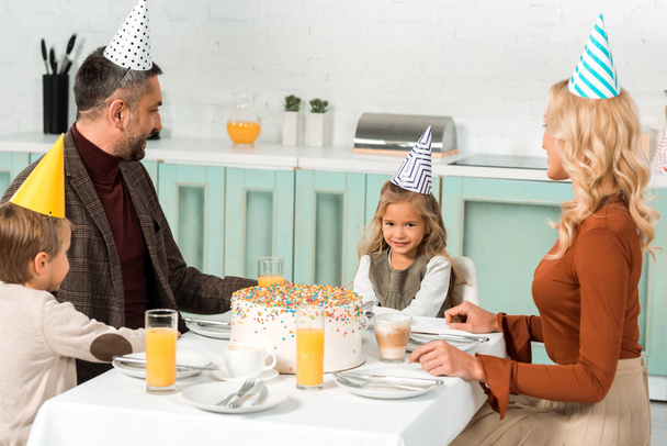 cute kid looking at birthday cake while sitting at served table near parents and brother - Photo, image