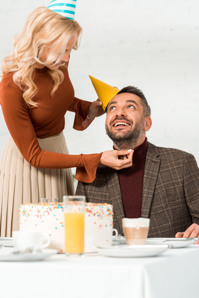 glimlachende vrouw zetten party cap op gelukkig echtgenoot zitten aan tafel met verjaardagstaart - Foto, afbeelding