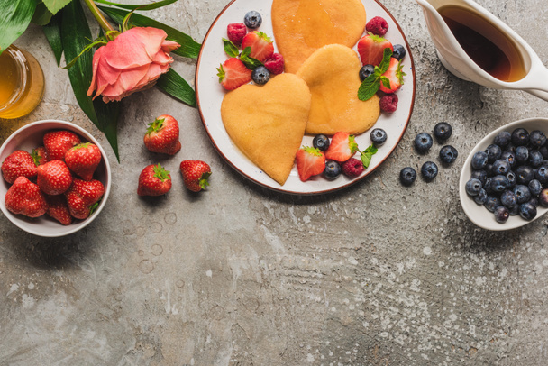 top view of heart shaped pancakes with berries on grey concrete surface with blooming rose, honey and maple syrup - Foto, Imagen