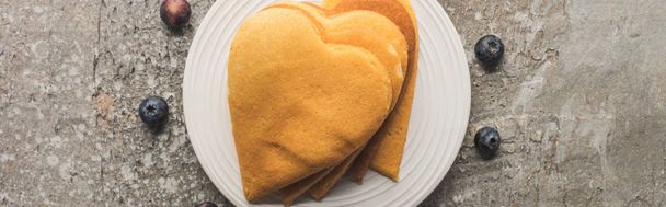 top view of delicious heart shaped pancakes on plate near blueberries on grey concrete surface, panoramic shot - Foto, imagen