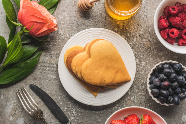 top view of heart shaped pancakes with berries, honey, cutlery and rose on grey concrete surface - Foto, Imagem