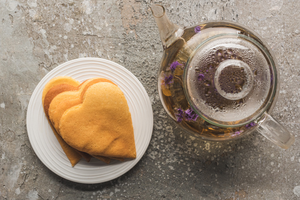 top view of heart shaped pancakes near herbal tea in teapot on grey concrete surface - Fotoğraf, Görsel