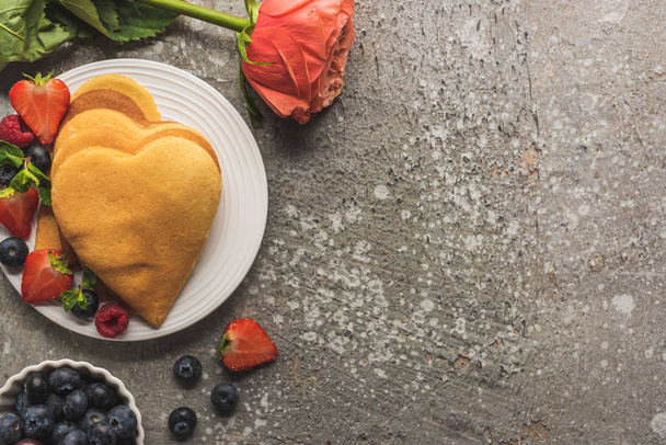 top view of heart shaped pancakes with fresh berries on grey concrete surface with rose - Фото, изображение