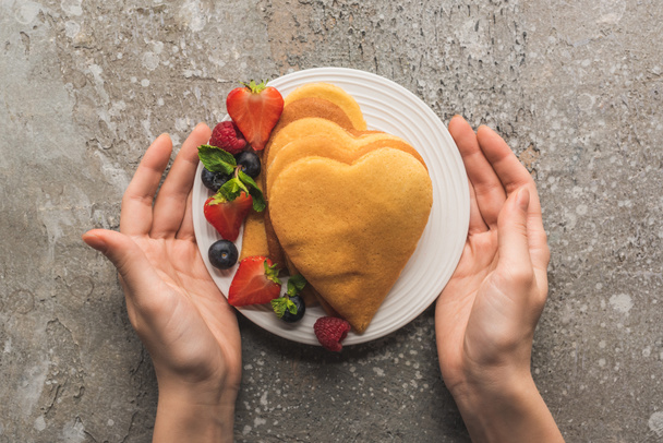 partial view of woman holding plate with heart shaped pancakes and tasty berries on grey concrete surface - Foto, immagini