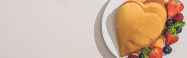 Draufsicht auf herzförmige Pfannkuchen mit leckeren Beeren auf Teller auf weißem Hintergrund, Panoramaaufnahme - Foto, Bild