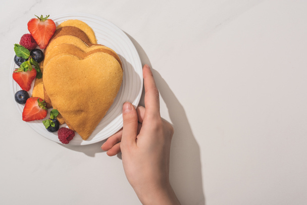 oříznutý pohled na ženu držící talíř s palačinkami ve tvaru srdce a chutnými bobulemi na talíři na bílém pozadí - Fotografie, Obrázek