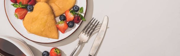 top view of delicious heart shaped pancakes with berries near cutlery and maple syrup on white background, panoramic shot - Foto, immagini