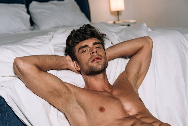 handsome, shirtless man looking at camera while lying on floor near bed - Fotó, kép
