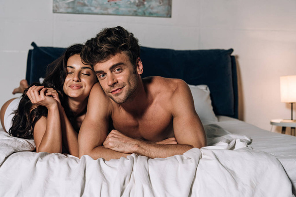 uomo allegro guardando la fotocamera mentre sdraiato sul letto con fidanzata felice
 - Foto, immagini