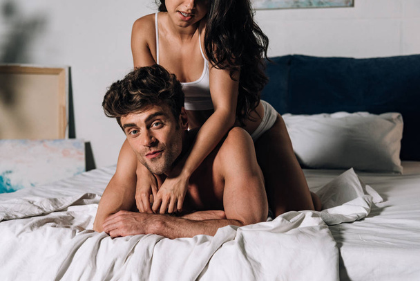 обрезанный вид сексуальной девушки, обнимающей мужчину, лежащего на кровати и смотрящего в камеру
 - Фото, изображение