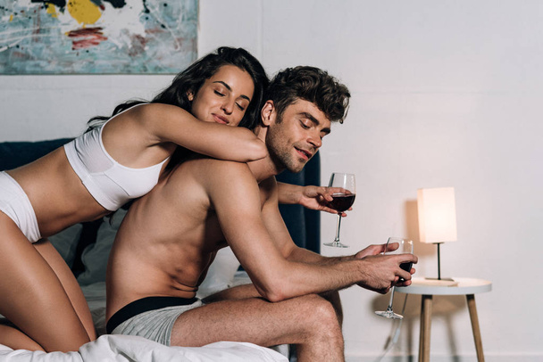 happy couple in underwear holding glasses of red wine while sitting on bed - Foto, immagini
