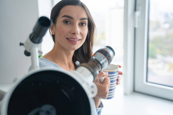 Smiling woman near the window with mug and telescope stock photo - Foto, Imagen