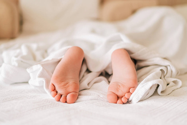 ベッドの上の白い毛布の下に小さな眠っている子供の足. - 写真・画像