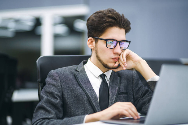 Бізнес-менеджер в окулярах працює на ноутбуці в сучасному офісі. Молодий працівник дивиться на монітор комп'ютера під час робочого дня в офісі. Молодий фрілансер, що працює з портативного комп'ютера
.  - Фото, зображення