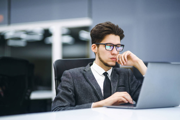 A szemüveges üzletvezető a laptopján dolgozik a modern irodában. Fiatal alkalmazott figyeli a számítógép monitort munka közben. Fiatal szabadúszó dolgozik egy laptop számítógép.  - Fotó, kép