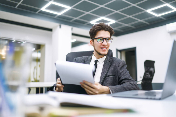 Young office worker in glasses reads documents in the modern office. Young business manager working with new startup project. Stylish man in a suit reviews and analyze documents, plans. - Photo, Image