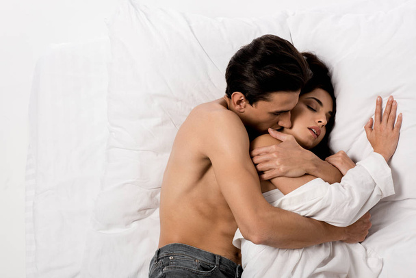 top view of tender boyfriend hugging sensual girlfriend and lying on white bed - Fotó, kép