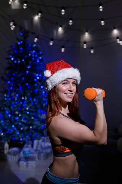 Beautiful redhead athlete in a Santa Claus hat is doing biceps exercises. Fitness woman in sportswear is holding dumbbells in her hands in a dark studio on the yon of the New Year tree. - Foto, afbeelding