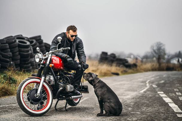 Een man met een baard in een zonnebril en een leren jas glimlachend op een rode motor op de weg met zijn hond. Achter hem is een rij banden. - Foto, afbeelding