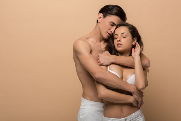 shirtless boyfriend hugging beautiful girlfriend in white bra, isolated on beige - Photo, Image