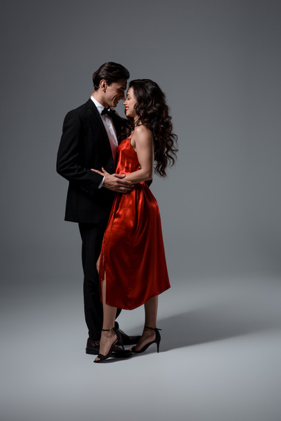Romantyczna para w garniturze i czerwonej jedwabnej sukience przytulająca się i patrząca na siebie na szaro - Zdjęcie, obraz
