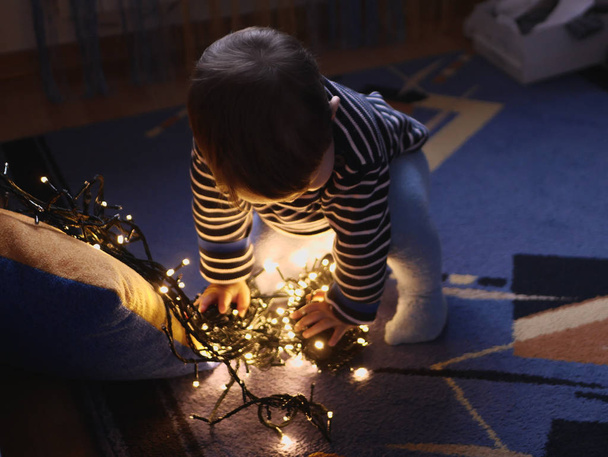 child and garland in the dark. little child holds shiny christmas lights indoors close dark background. Portrait of a happy kid boy smiling to a festive garland on the eve of the holiday of hands. The - Valokuva, kuva