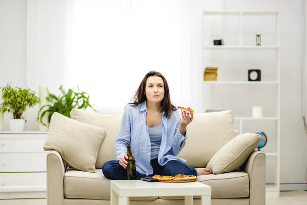 Hungry woman chews, holding a bottle of beer and a slice of pizza hands. Isolated woman on blurred light background. Copy space. - Foto, imagen