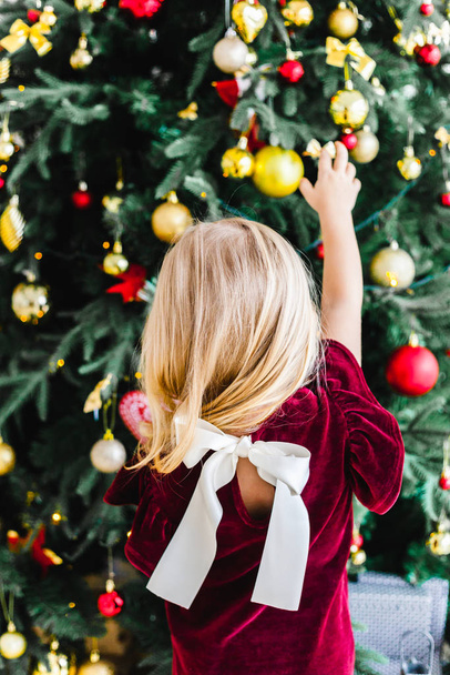A little girl in a Burgundy dress with a bow stands near the Christmas tree, waiting for the new year and Christmas - Фото, зображення