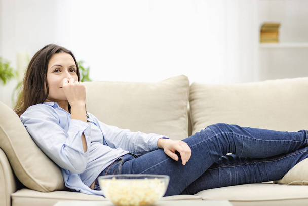 Portrait of pretty, charming, cheerful woman with straight black hair, who is eating pop corn, watching comic serial, comedy, sitting with crossed legs in living room. - Foto, imagen