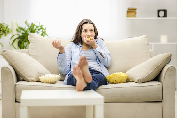 Portrait of pretty, charming, cheerful woman with straight black hair, who is eating pop corn and potato chips, watching comic serial, comedy, sitting with crossed legs in living room. - Fotó, kép