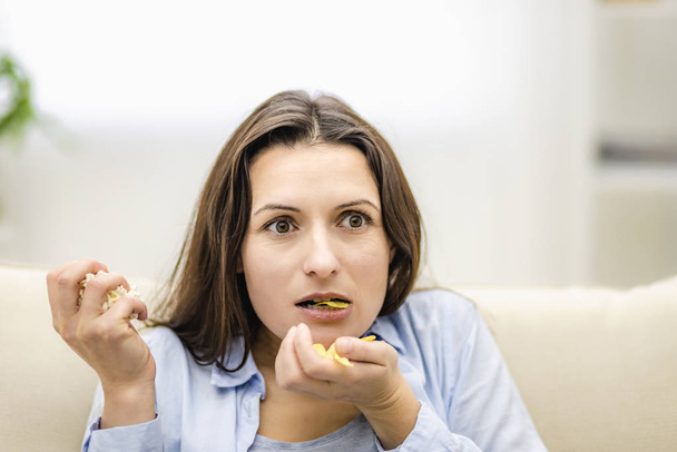 Frau reißt erstaunt den Mund auf. Sie hält zwei Handvoll Snacks in der Hand: Kartoffelchips und Popcorn. Nahaufnahme. - Foto, Bild