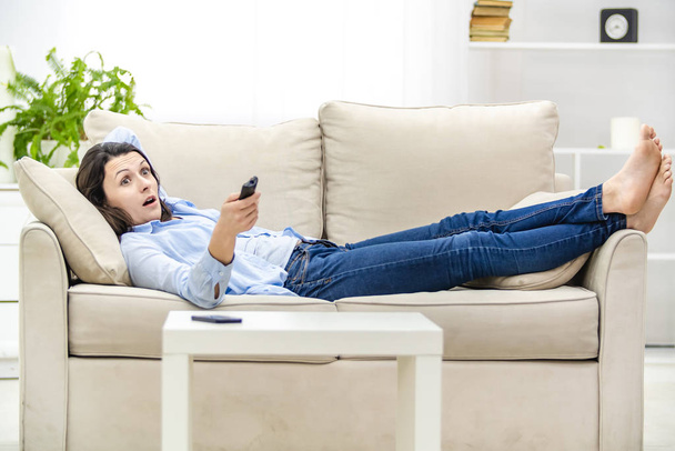 Shocked and surprised woman opens her mouth, while watching TV, laying on a sofa. Copy space. - Foto, Imagem