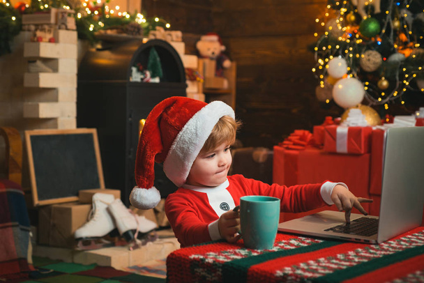 On-line jouluostoksia lapsille. Pikkupojalla on joulupukin vaatteet kannettavan vieressä. Joulupukin apulainen muistikirjan avulla. - Valokuva, kuva