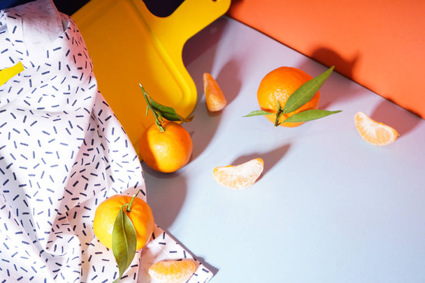 Mandarinas naranjas frescas con hojas con una tabla de cortar amarilla y una toalla, sobre un fondo colorido
 - Foto, Imagen
