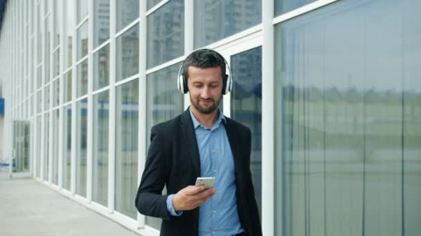 Relaxed businessman in headphones walking outdoors using smartphone smiling - Záběry, video